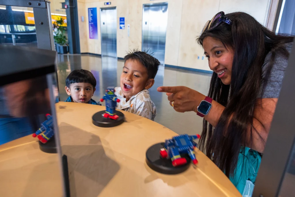 A woman and two young boys are excitedly interacting with small, colorful robot models on Dancing Robots exhibit. The woman points at one robot while the boys watch with amazement and curiosity.