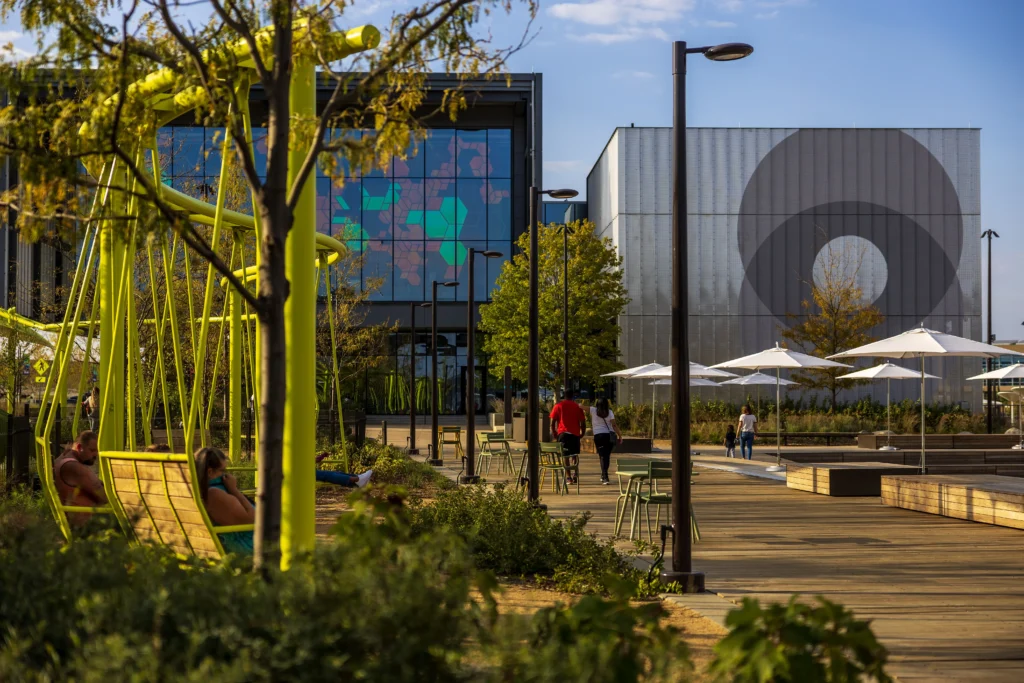 Exterior view of the Luminarium from the Heartland of America Park featuring bright yellow swings, greenery, walkways, tables, and people strolling.